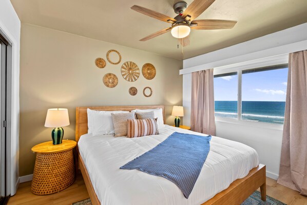 A bedroom with a bed, ceiling fan, decorative wall art, bedside lamps, and a view of the ocean. - The Beach Bungalow | Ocean Front (San Diego)