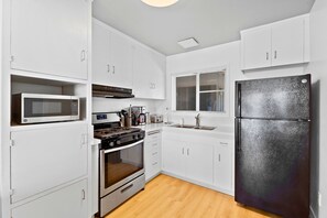 A kitchen with white cabinets, a black refrigerator, a microwave, a stove, and a window with blinds. - The Beach Bungalow | Ocean Front (San Diego)