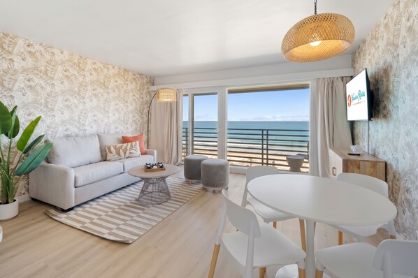 A modern living room with a sofa, coffee table, and dining area, overlooking a beach. - Sea Cliff #8 | La Palma (San Diego)