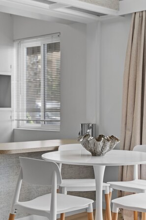 A modern dining area with white round tables and chairs, a decorative bowl on one table, and a window with blinds. - Seabreeze - Ocean Front (San Diego)