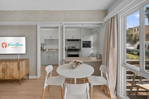 A modern kitchen with white cabinets, a dining area with a white table and chairs, and a large window with curtains. - Seabreeze - Ocean Front (San Diego)