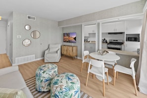 A modern living room with a sofa, ottomans, a dining table with chairs, a TV, and a kitchen area with cabinets and appliances. - Seabreeze - Ocean Front (San Diego)