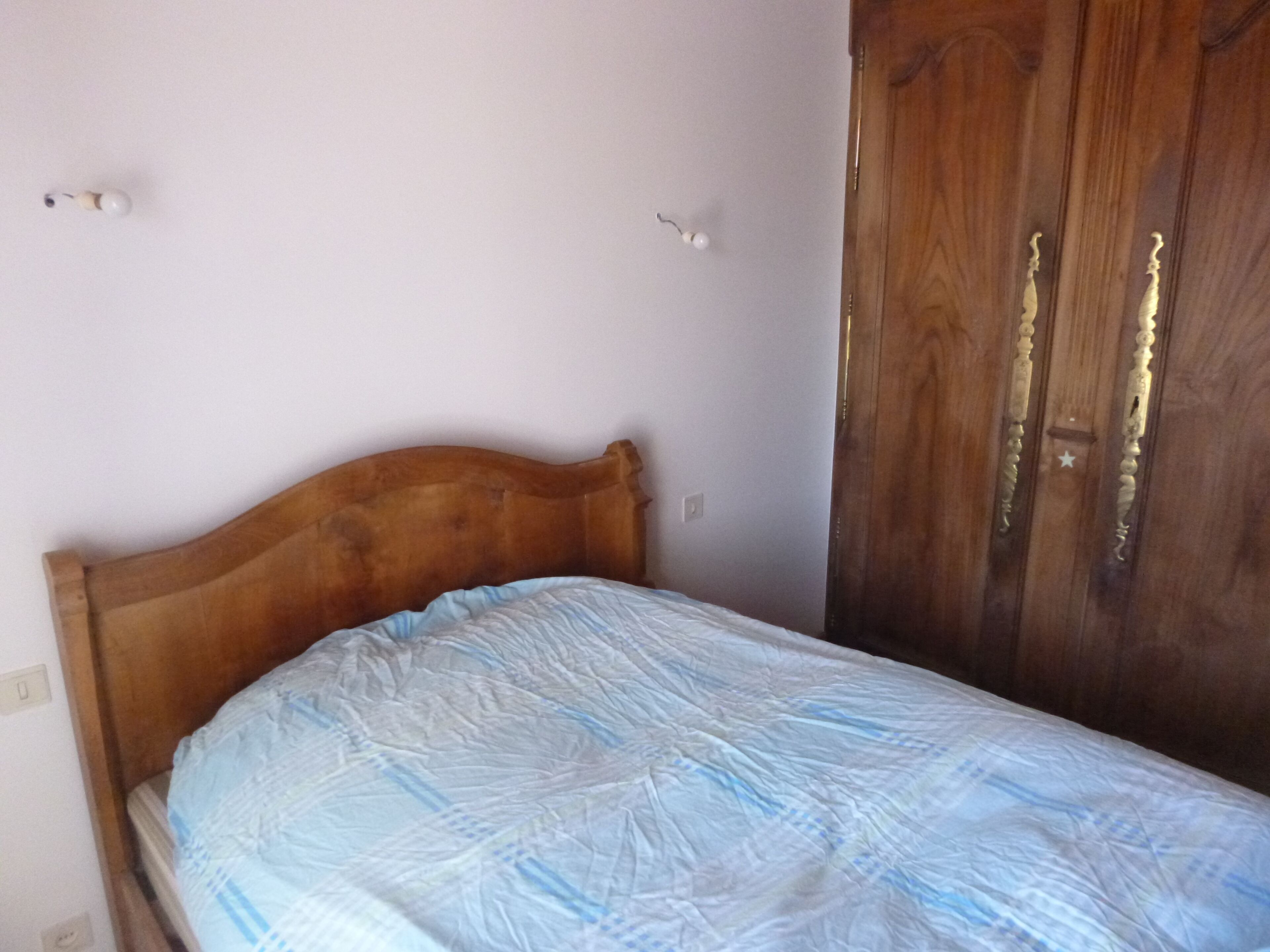 Un lit simple avec une tête de lit en bois, dans une pièce aux murs de couleur claire et dotée d’une armoire en bois.
