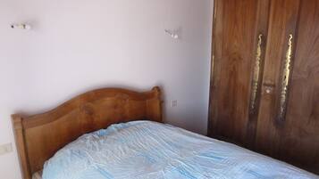 Un lit simple avec une tête de lit en bois, dans une pièce aux murs de couleur claire et dotée d’une armoire en bois.