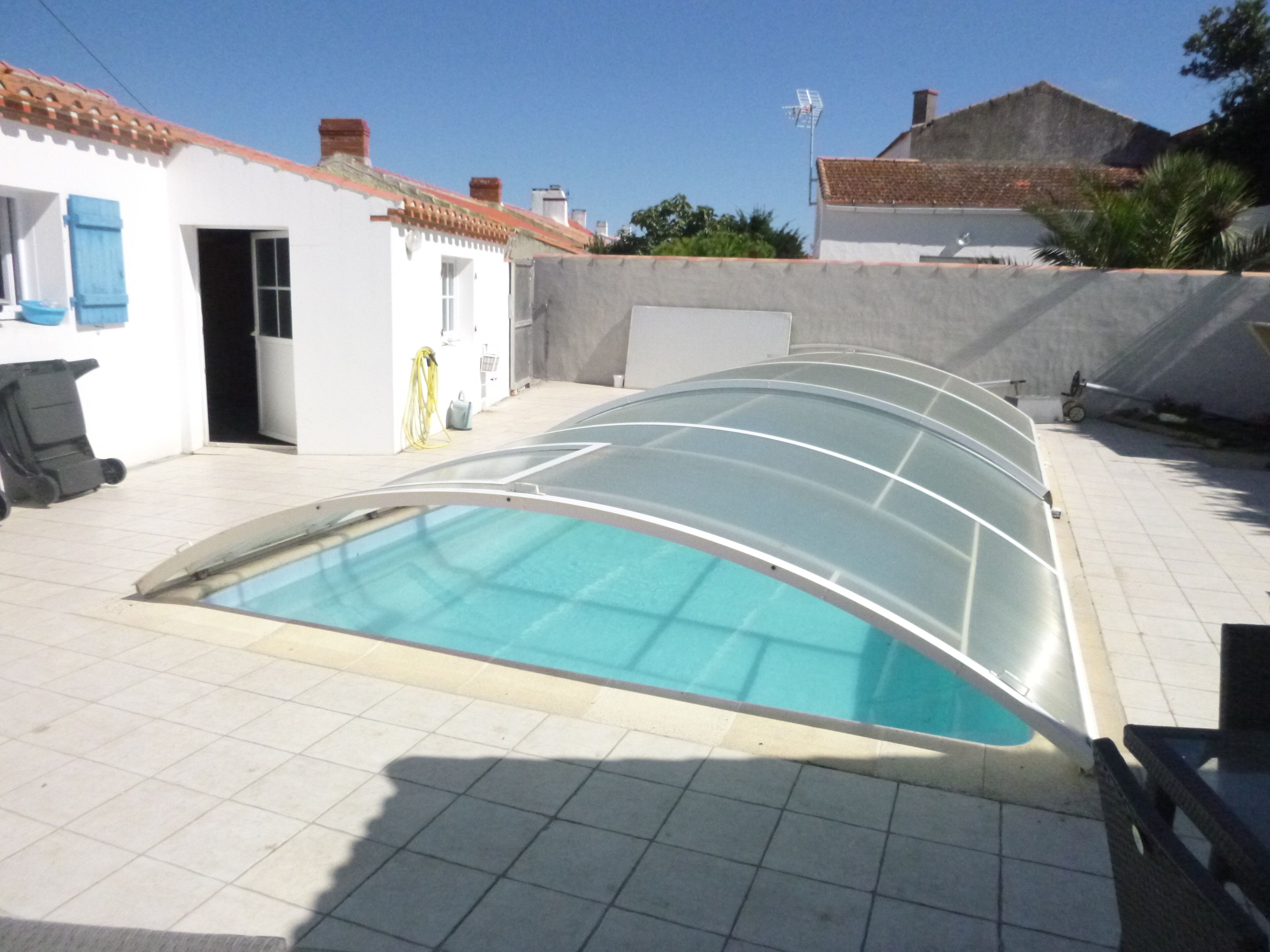 Une piscine entourée de parois en verre, un bâtiment blanc avec un toit recouvert de tuiles et un ciel dégagé.