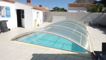 Une piscine entourée de parois en verre, un bâtiment blanc avec un toit recouvert de tuiles et un ciel dégagé.