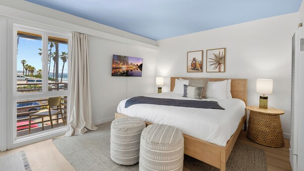 Ein Hotelzimmer mit Bett, zwei Hocker, Blick auf Palmen und Stadtansicht sowie ein Balkon mit einem Stuhl.
