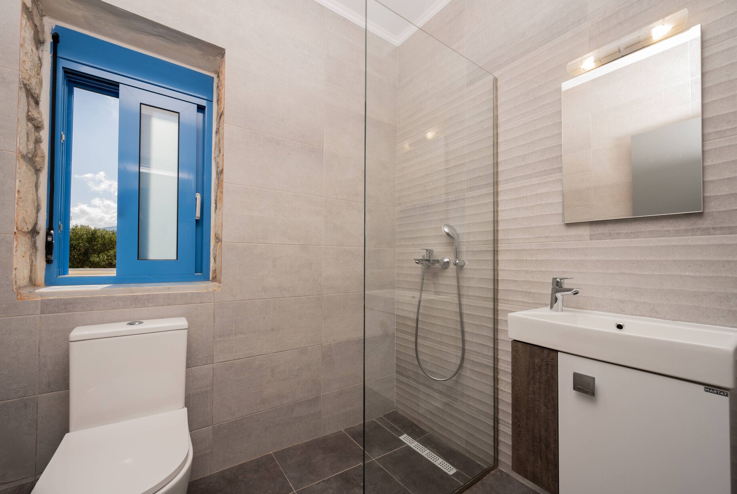 Un baño moderno con ducha de vidrio, inodoro blanco, lavamanos y ventana con marco azul.