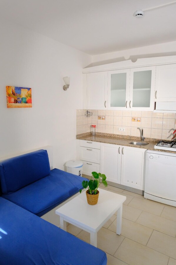 A compact kitchen with white cabinets, a sink, and a dishwasher. There's a blue sofa and a small table with a plant. - Aktur Cove Apart Hotel (Bodrum)