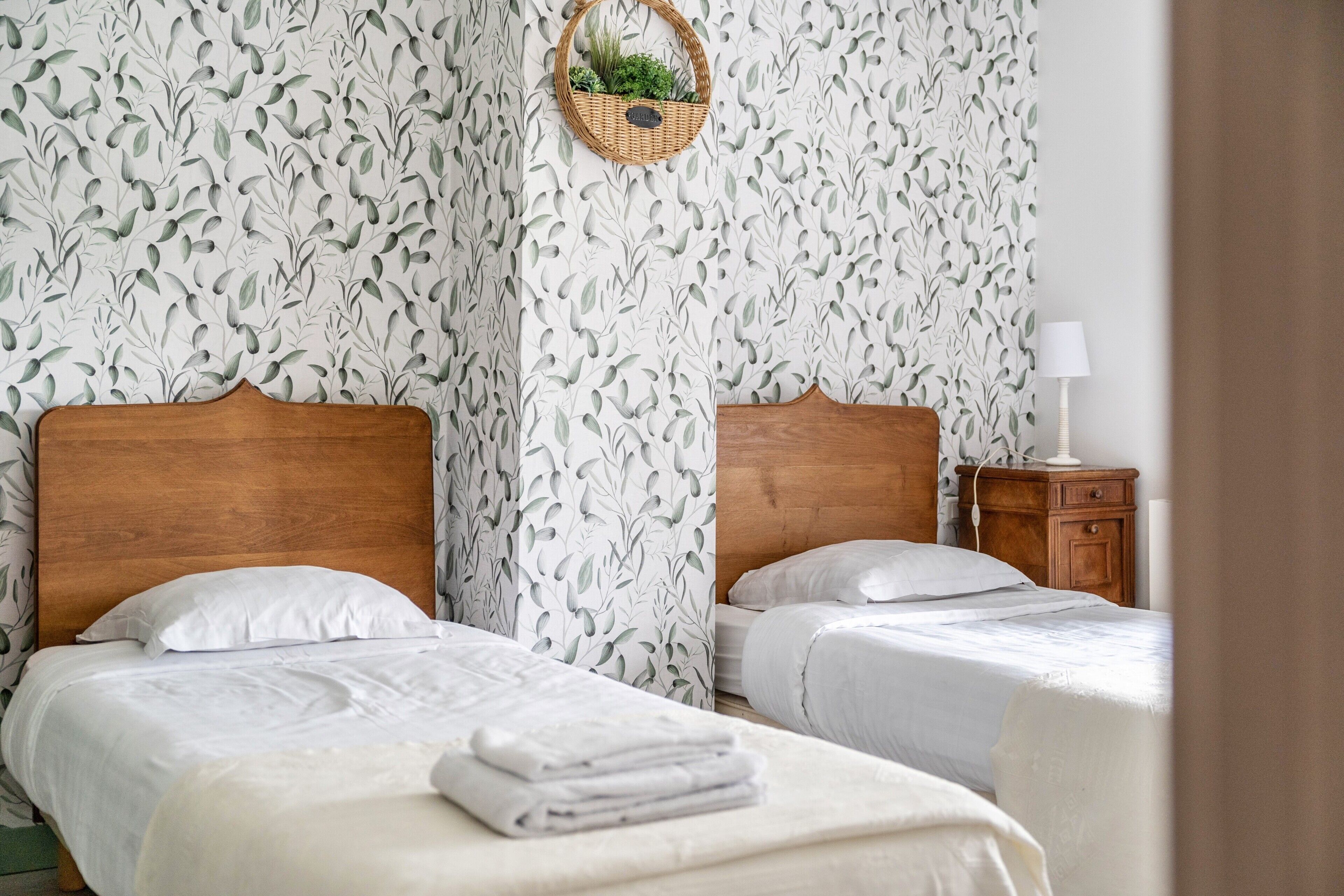 A hotel room with two beds, a wooden headboard, a nightstand, a lamp, and a wall with a leafy pattern.