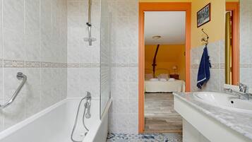 Een badkamer met een bad, douche en wastafel, met een gang die leidt naar een slaapkamer.