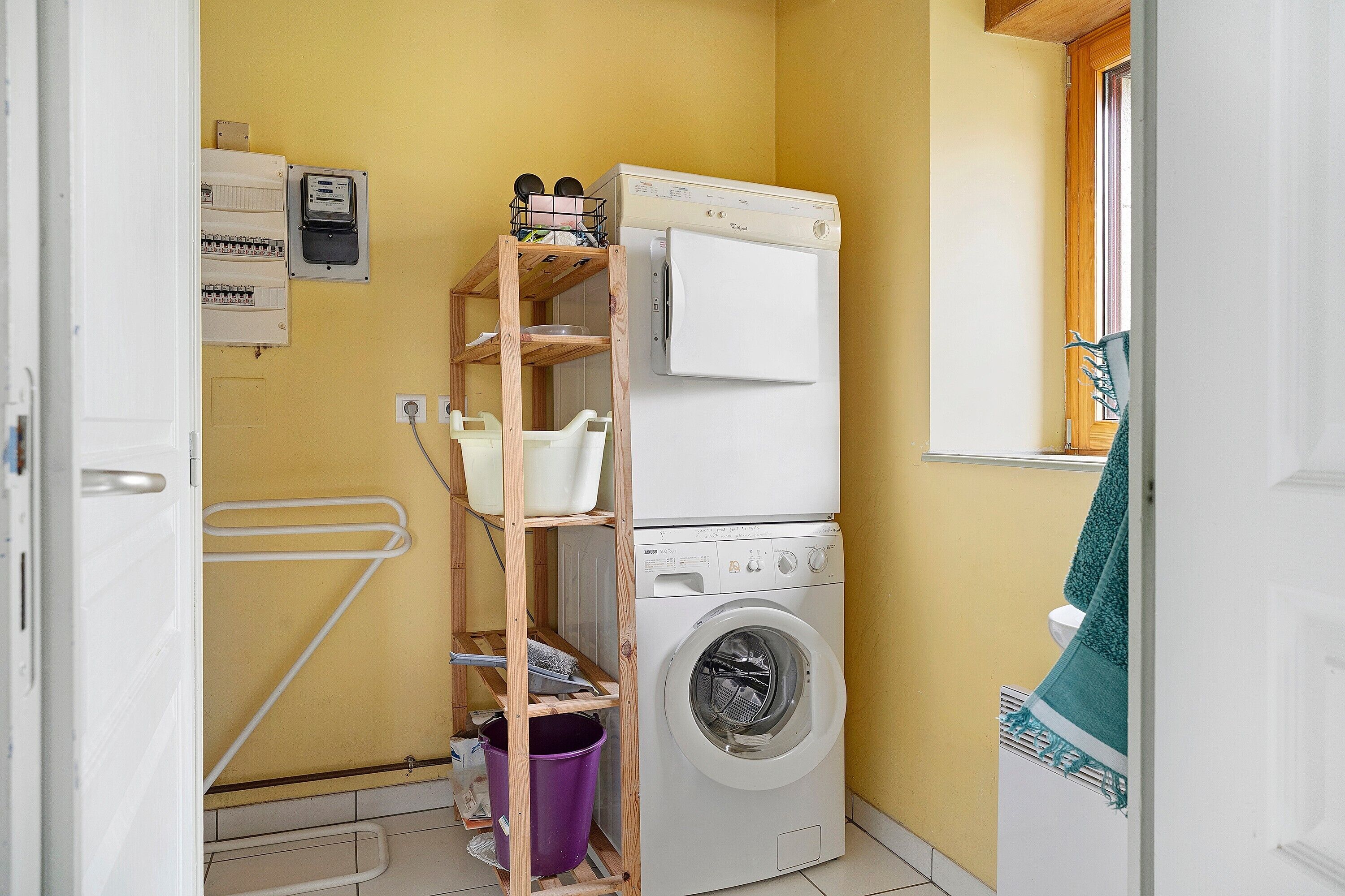 Une petite buanderie équipée d’un lave-linge, d’un sèche-linge et d’une étagère en bois où sont rangés divers objets.