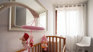 Ein Babybett mit Mobile, einem Spiegel und einem weißen Stuhl in einem Zimmer mit Fenster und Vorhängen.