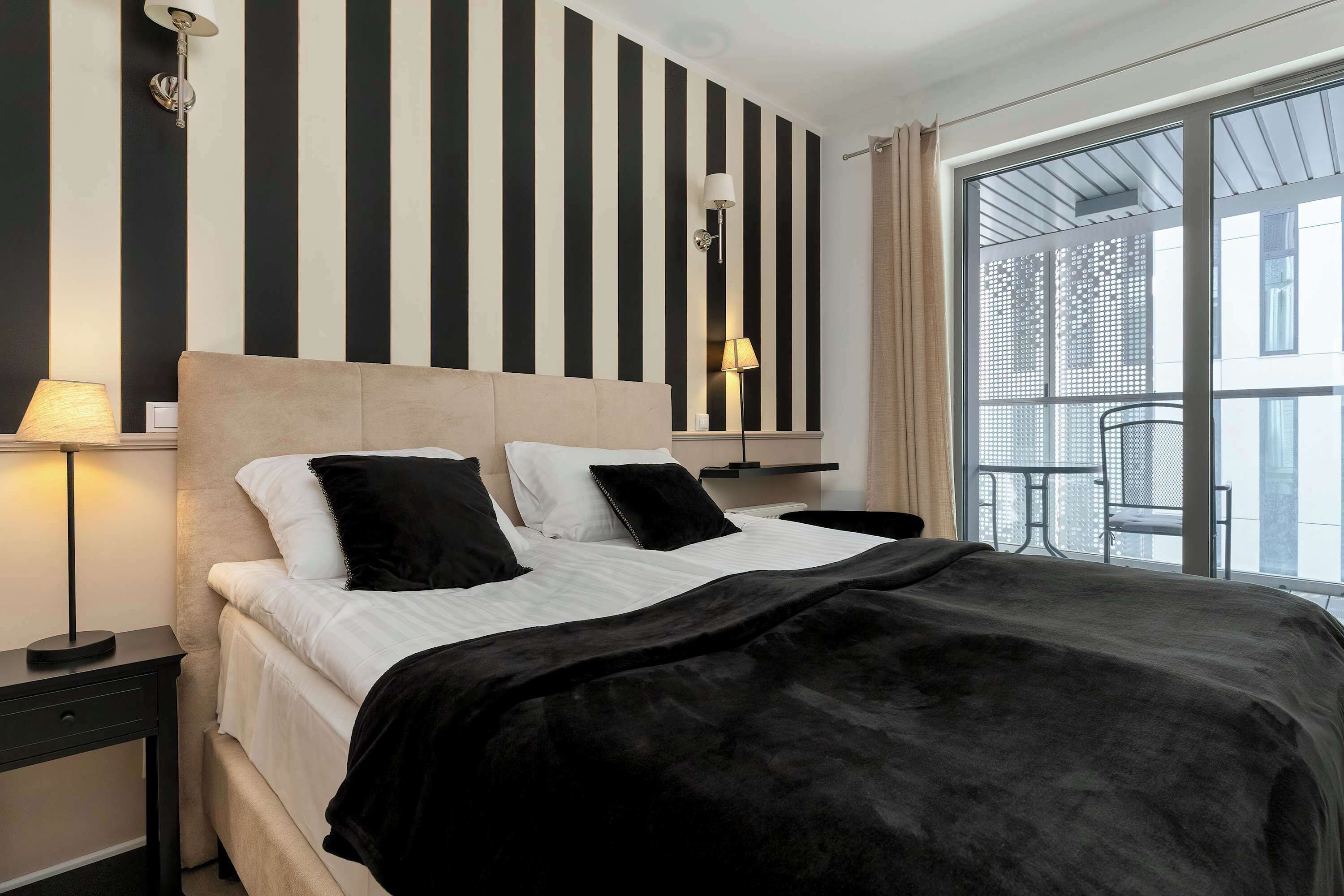 A hotel room with a bed, a nightstand, a lamp, a window with a view, and a wall with vertical stripes.
