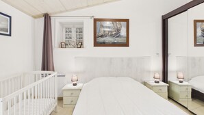 Un dormitorio con una cuna, una cama, dos mesitas de noche con lámparas, una ventana con cortinas y un cuadro en la pared.