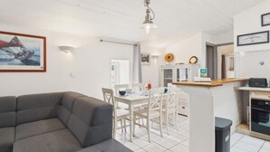 Una sala de estar con sofá gris, una zona de comedor con sillas blancas, una cocina con mostrador de madera y un ventilador de techo.
