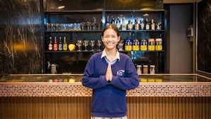 A person standing at a bar with shelves stocked with bottles and glasses behind them. - Queen On Street Guesthouse (Phuket)