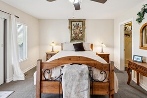 Een slaapkamer met een houten bed, twee nachtkastjes met lampen, een plafondventilator, een lijst met bloemkunst en een spiegel aan de muur.
