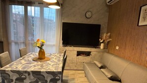 Een woonkamer met een bank, een eettafel met een tafelkleed met dessin, een wandtelevisie en een klok.