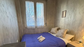 Een slaapkamer met een bed, kussens, een nachtkastje, een raam met gordijnen en een aan de muur hangend schilderij.