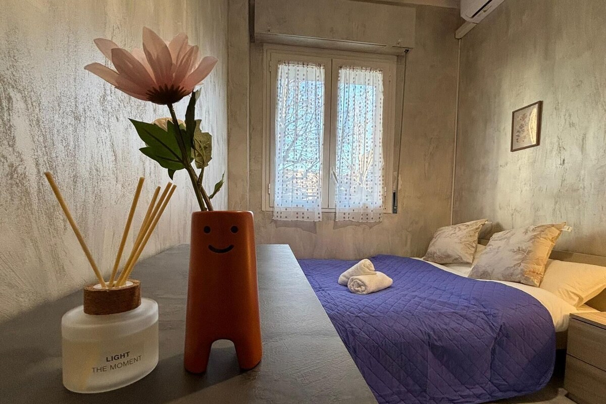 Un dormitorio con cama, ventana con cortinas, un florero con una flor y una botella etiquetada 'LIGHT THE MOMENT'.