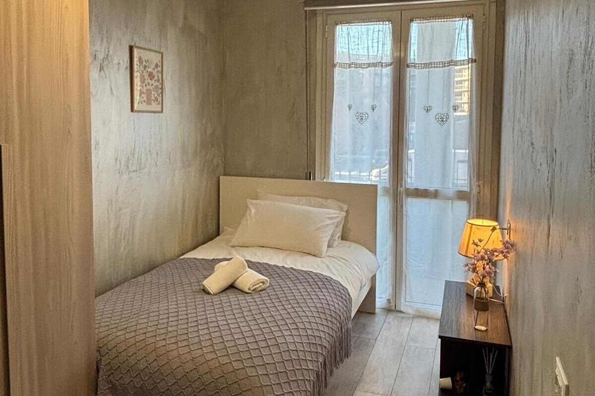 Un dormitorio con cama, mesita de noche con lámpara, un cuadro en la pared y ventana con cortinas.
