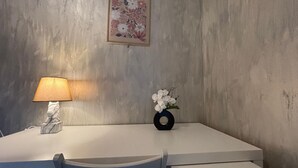 Ein Zimmer mit Schreibtisch, Lampe, Blumen in einer Vase und einem floralen Bild in einem Rahmen an der Wand.