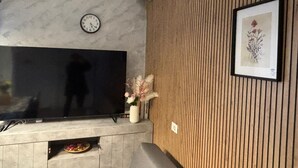 Een woonkamer met een flatscreen televisie, een klok, een lijst met afbeelding en een vaas met bloemen.