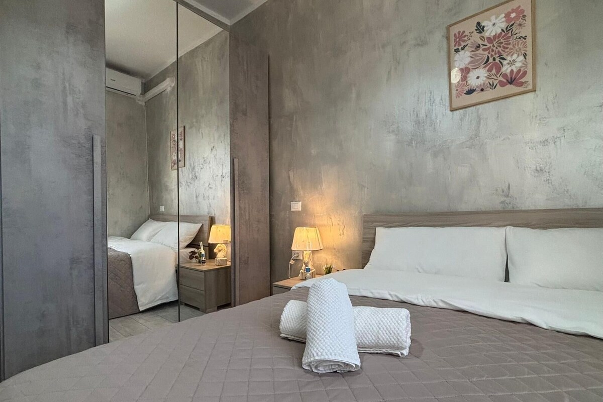 Habitación de hotel con una cama grande, una mesita de noche con lámpara, un espejo y una obra de arte floral en la pared.