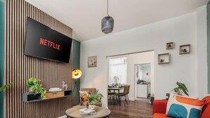 Moderni olohuone, jossa seinälle on asennettu televisio, jossa näkyy Netflixin logo. Olohuoneessa on myös ruokailutila tuoleineen ja sohvapöytä. Avonaisen oven läpi näkyy keittiö.