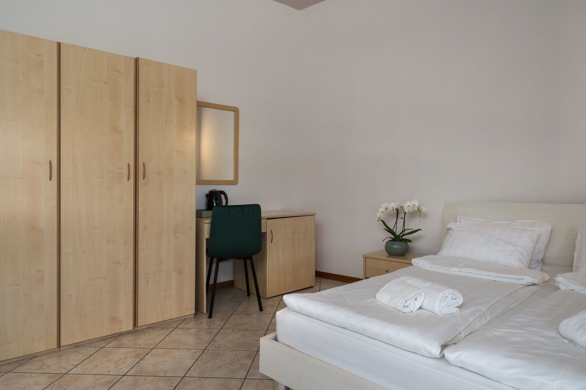 A hotel room with a bed, a desk, a chair, a wardrobe, and a nightstand with a vase of flowers.