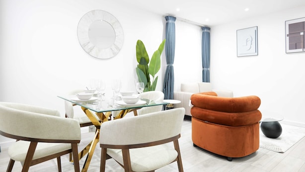 A modern dining area with a glass table, white chairs, and a large plant. - The Wembley Crown Luxe 1 Bed Flat  (Wembley)