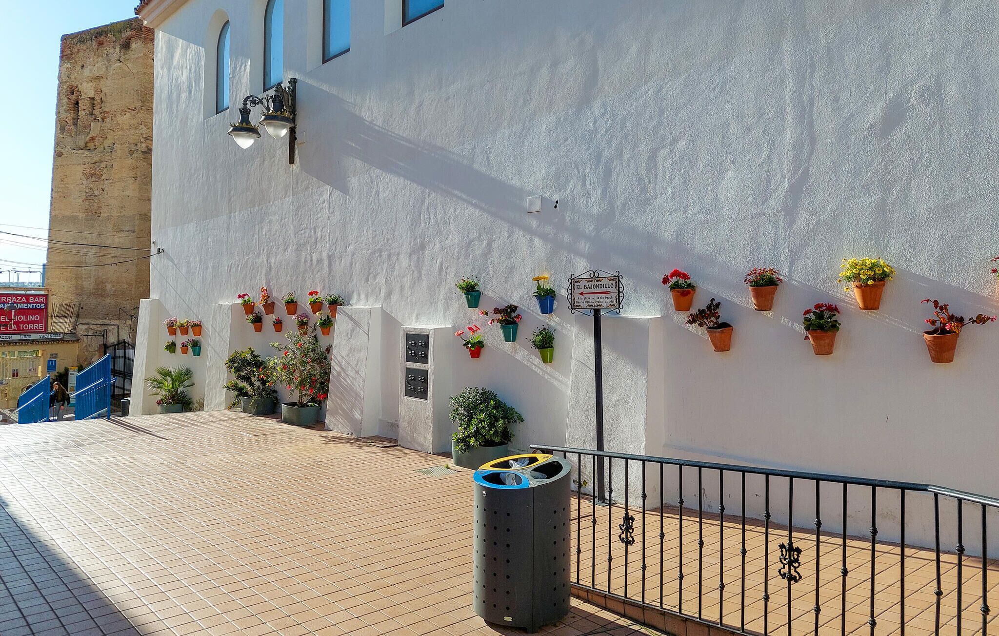 A building with a white facade, outdoor potted plants, a trash bin, and a sign indicating 'El Bajondillo.'