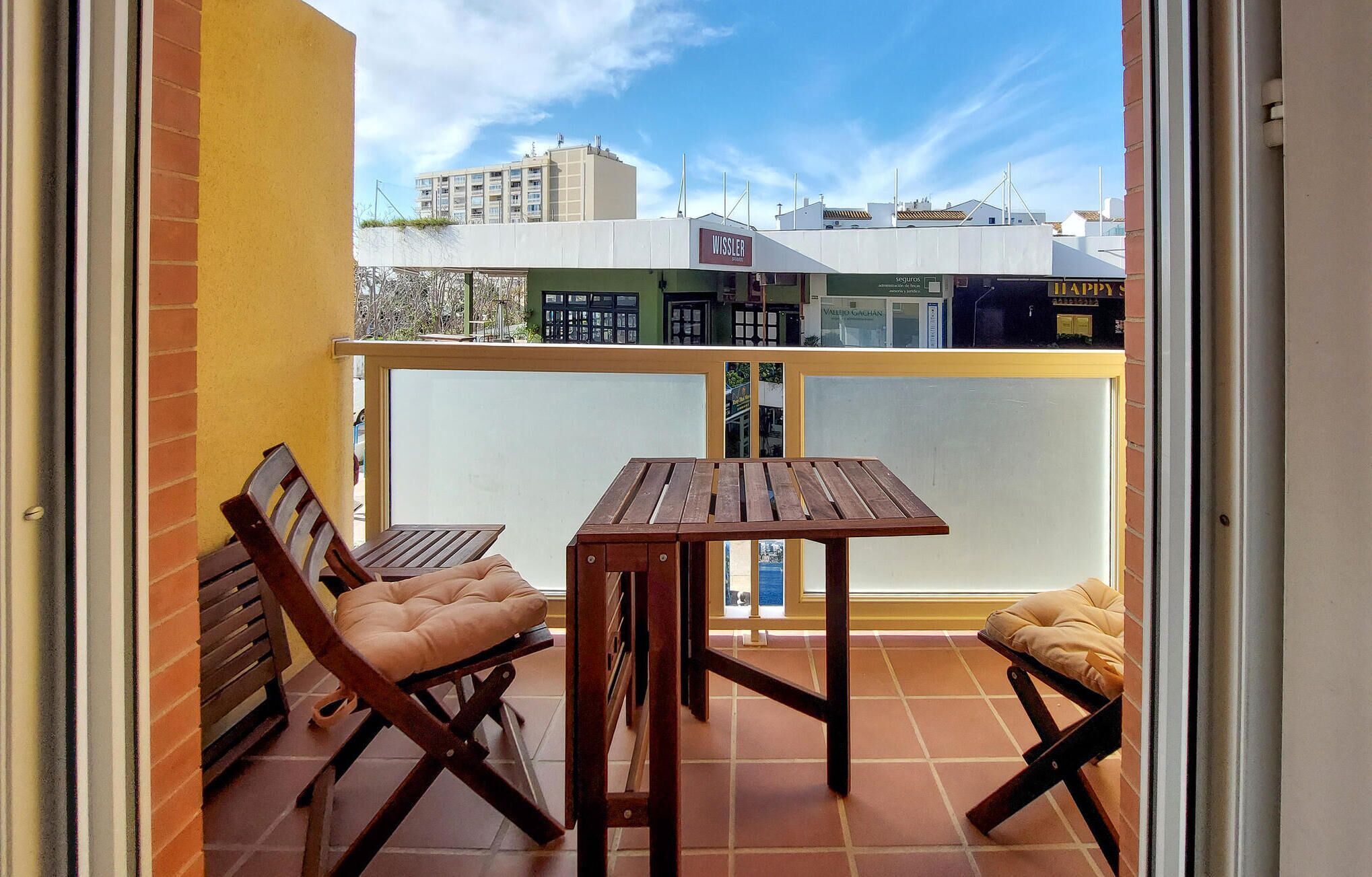 A balcony with two wooden chairs and a table, overlooking a cityscape with buildings and a clear sky.