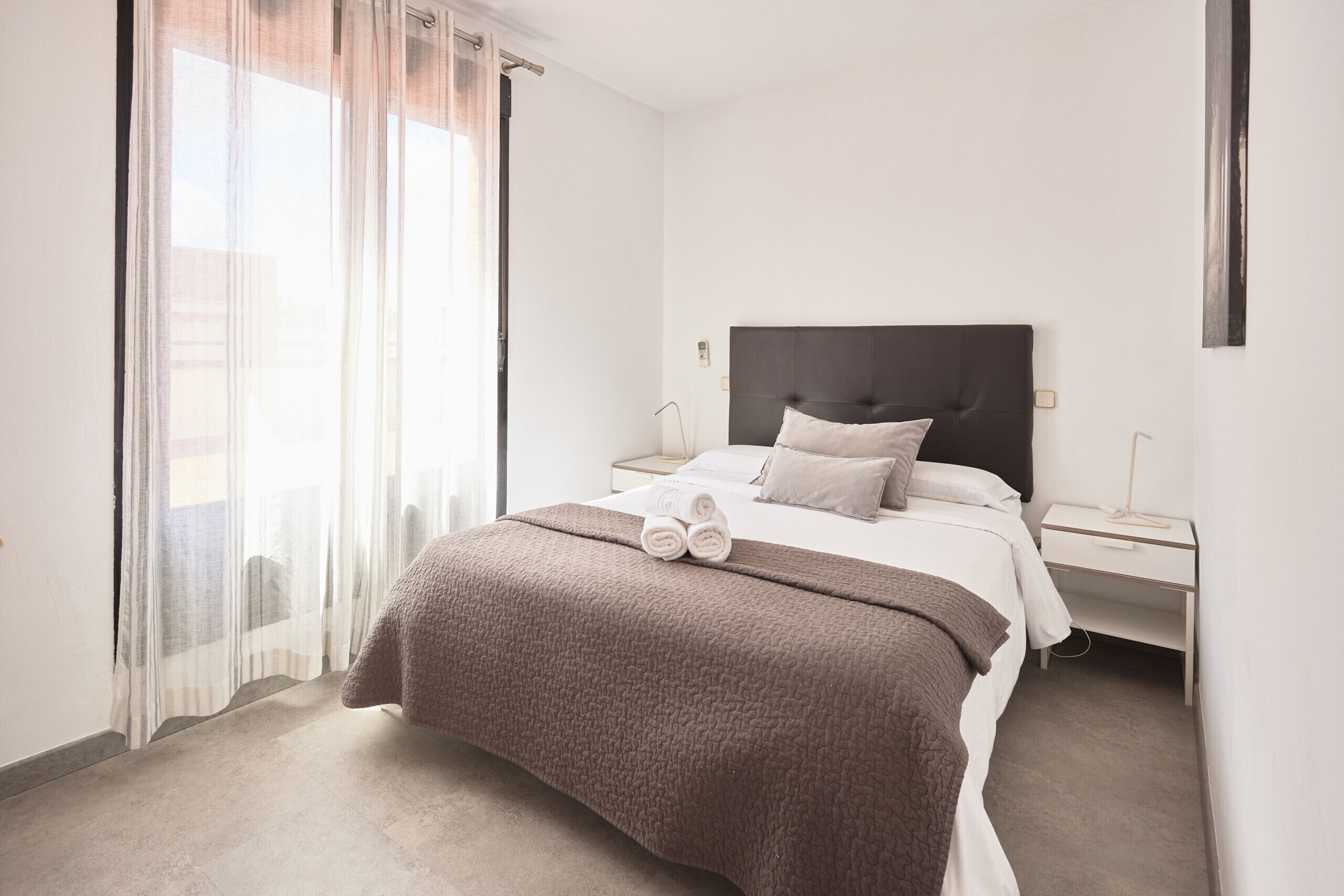A hotel room with a bed, a nightstand, a window with sheer curtains, and a view of the outside.