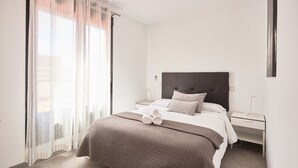 Hotellihuone, jossa on sänky, yöpöytä, ikkuna, jossa on läpikuultavat verhot, ja näkymä ulos.