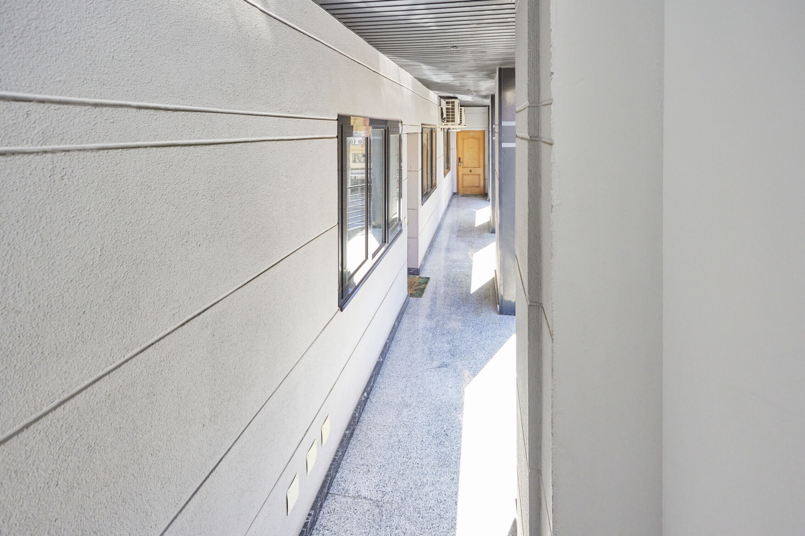 A narrow corridor with white walls, large windows, and a door at the end.