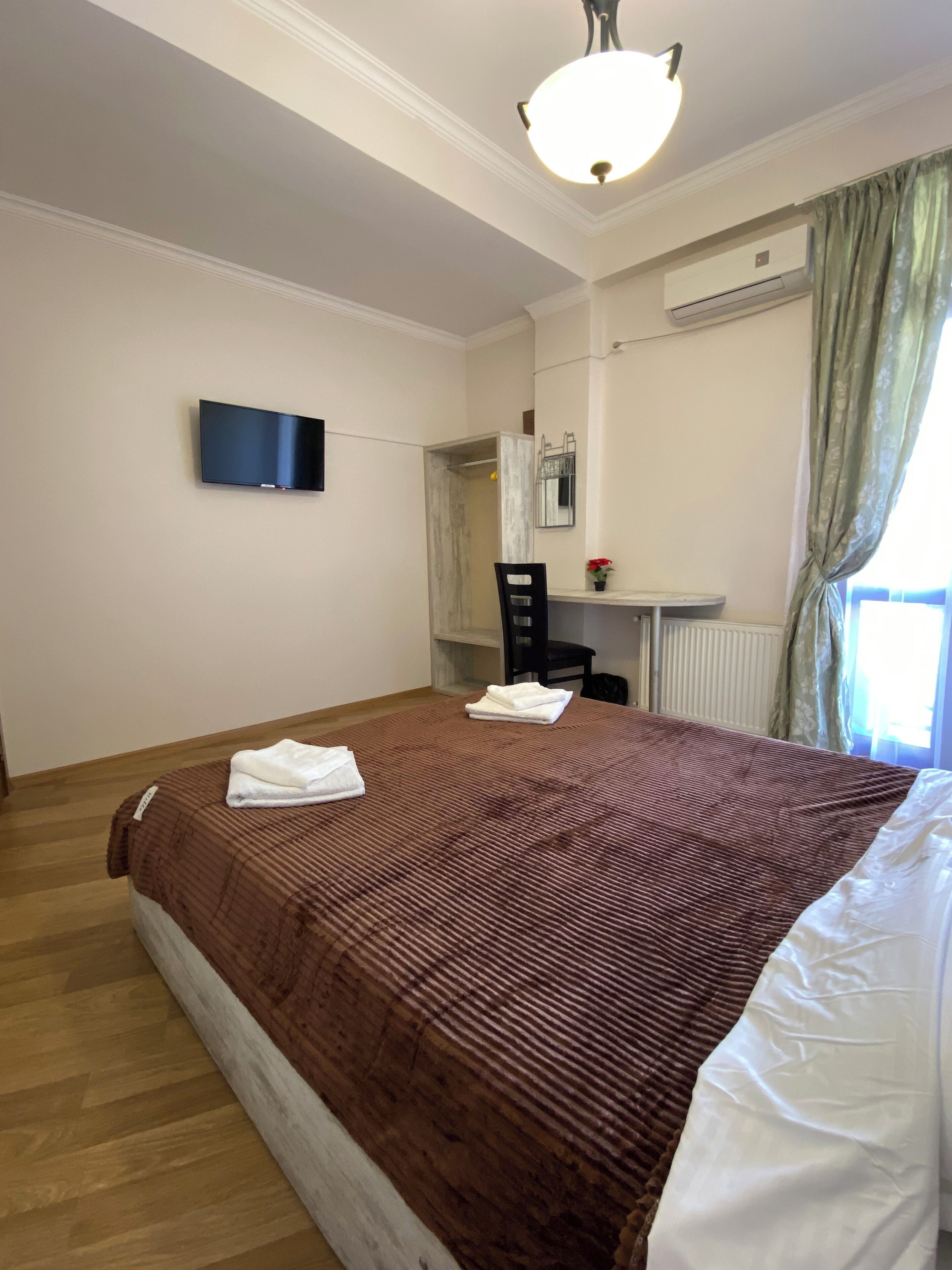 A hotel room with a bed, a flat-screen TV, a desk with a chair, a window with curtains, and an air conditioning unit.
