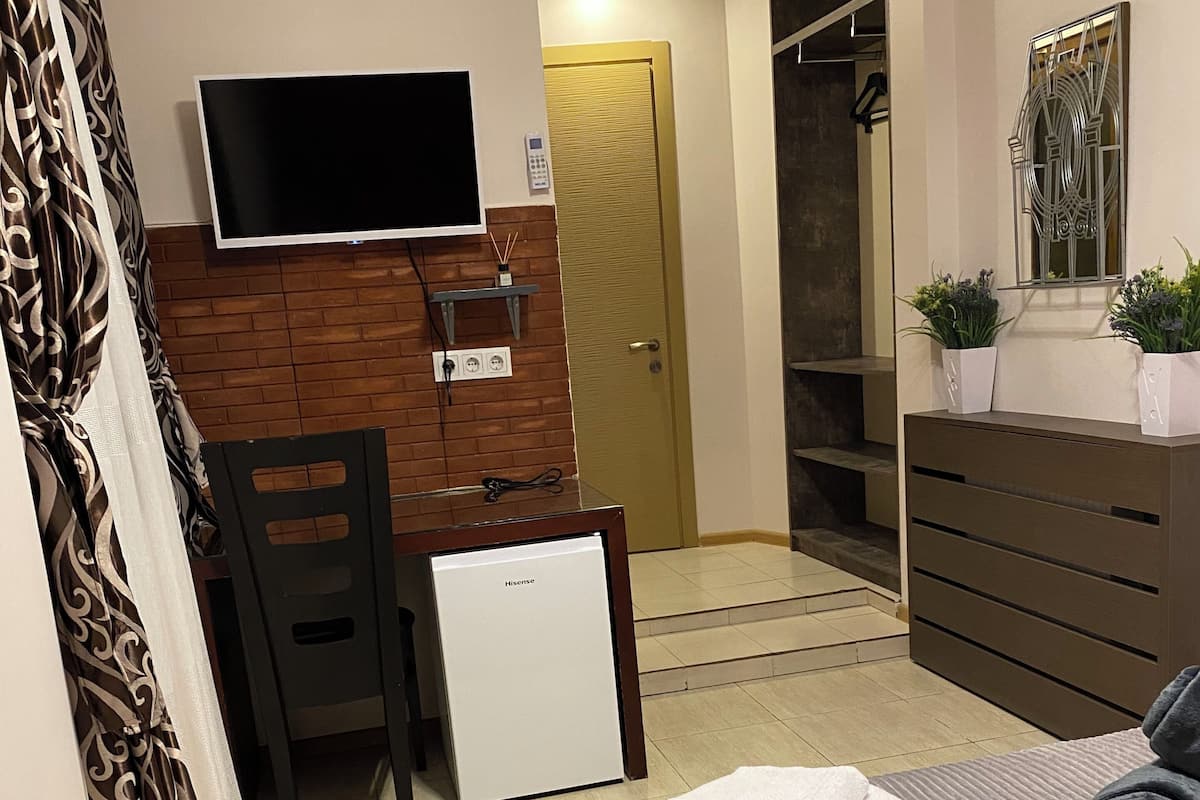 A hotel room with a bed, a TV mounted on the wall, a desk with a chair, a wardrobe, and a door leading to a bathroom.