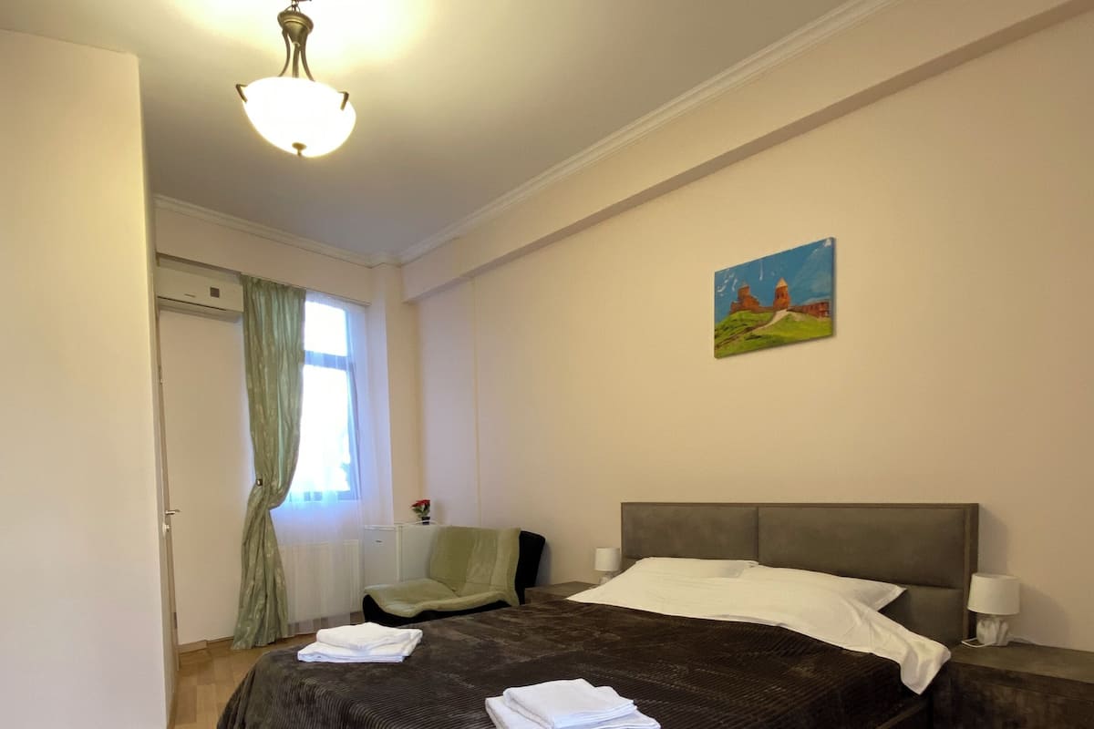 A bedroom with a large bed, a bedside table, a chair, a painting on the wall, and a window with curtains.