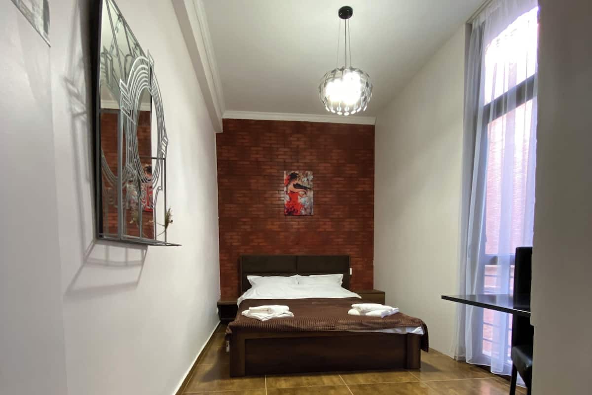 A bedroom with a bed, a mirror, a painting, a ceiling light, and a window with curtains.