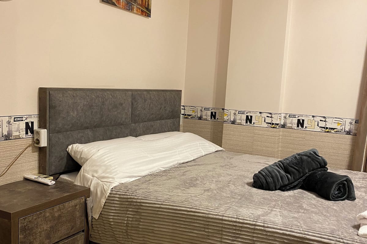 A bedroom with a bed, a nightstand, a painting, and an air conditioning unit.