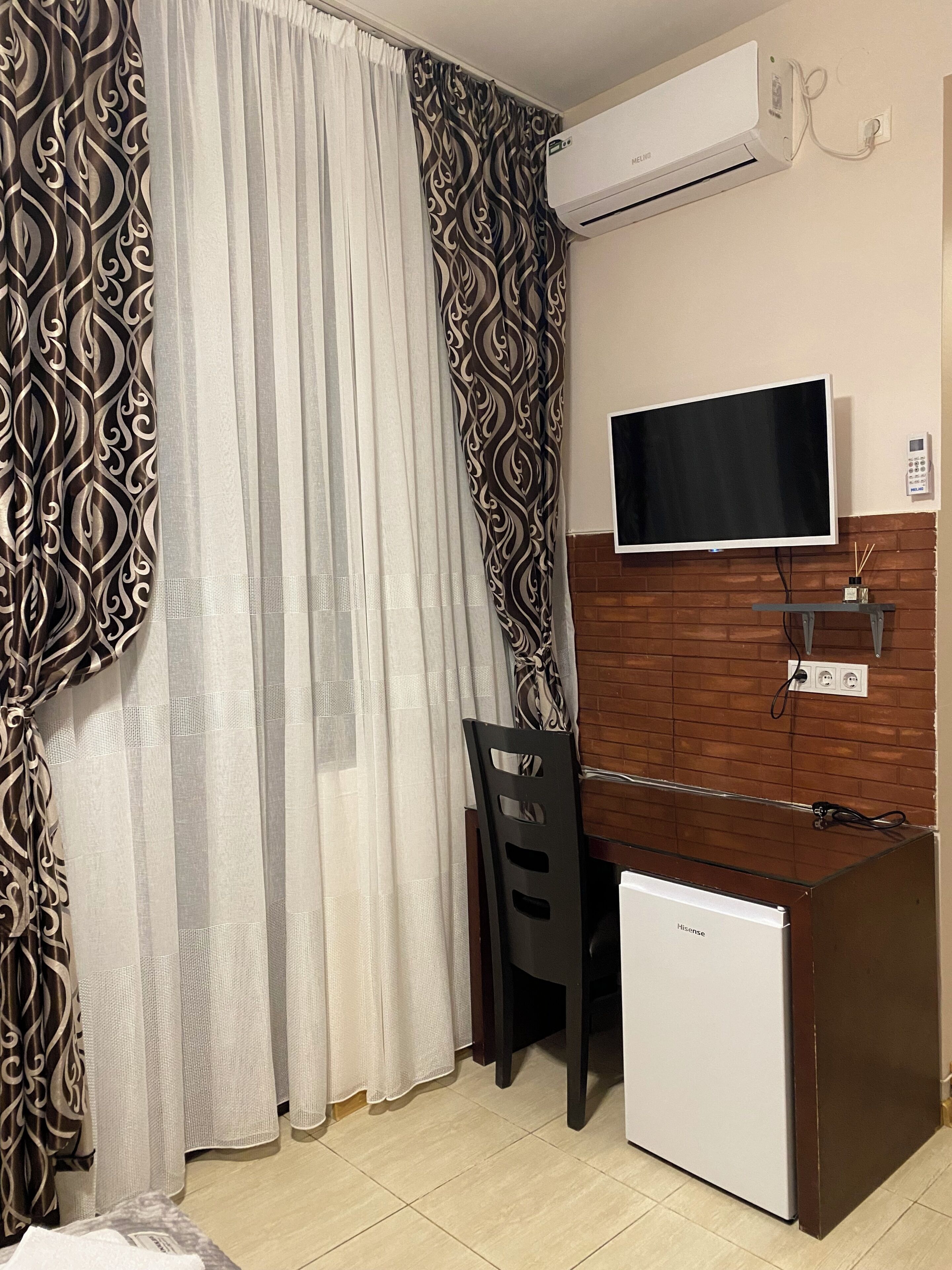 A room with a bed, a desk with a chair, an air conditioner, a television, and a small refrigerator.