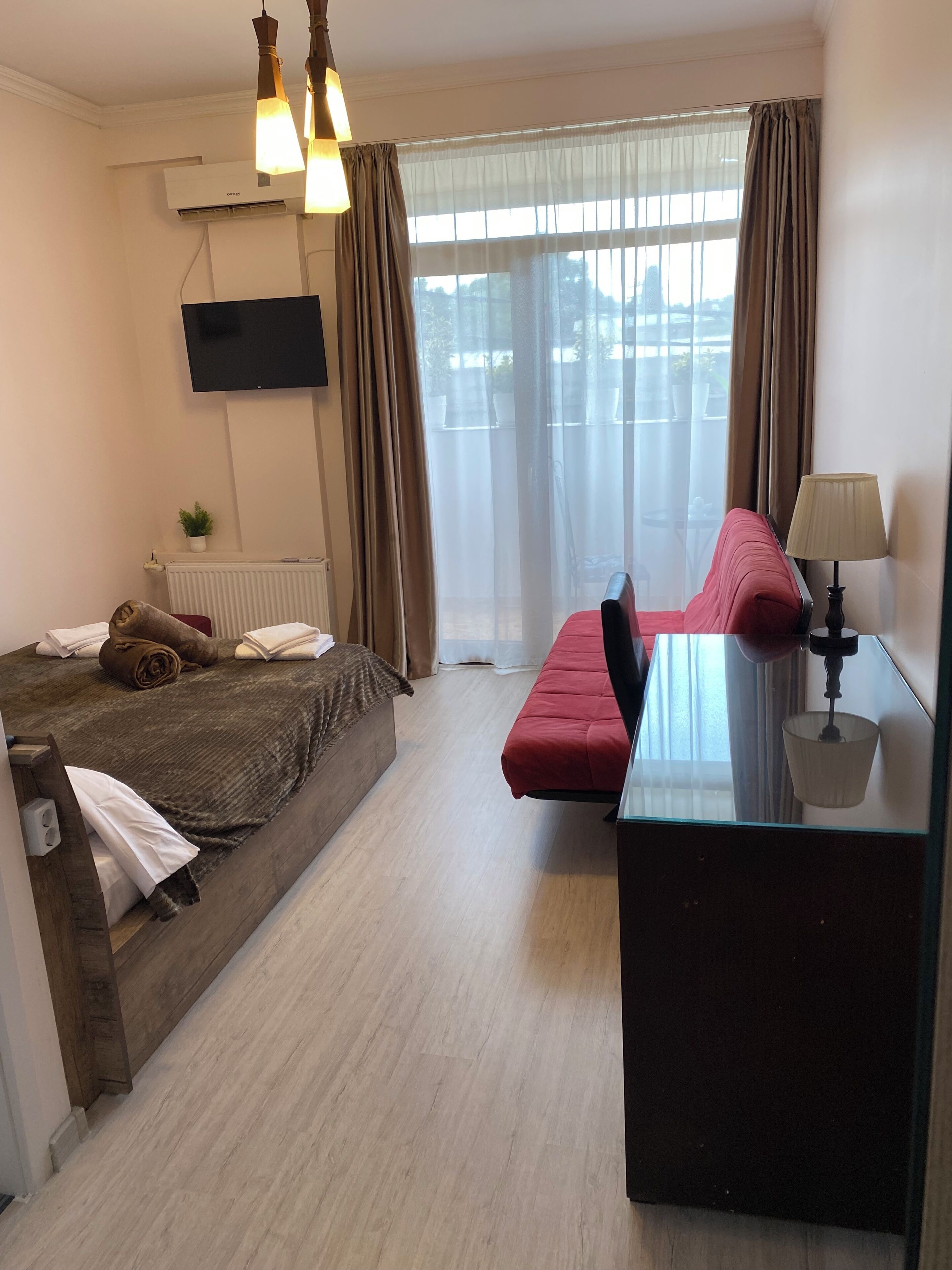 A hotel room with a bed, a sofa, a desk with a lamp, a TV mounted on the wall, and a window with curtains.