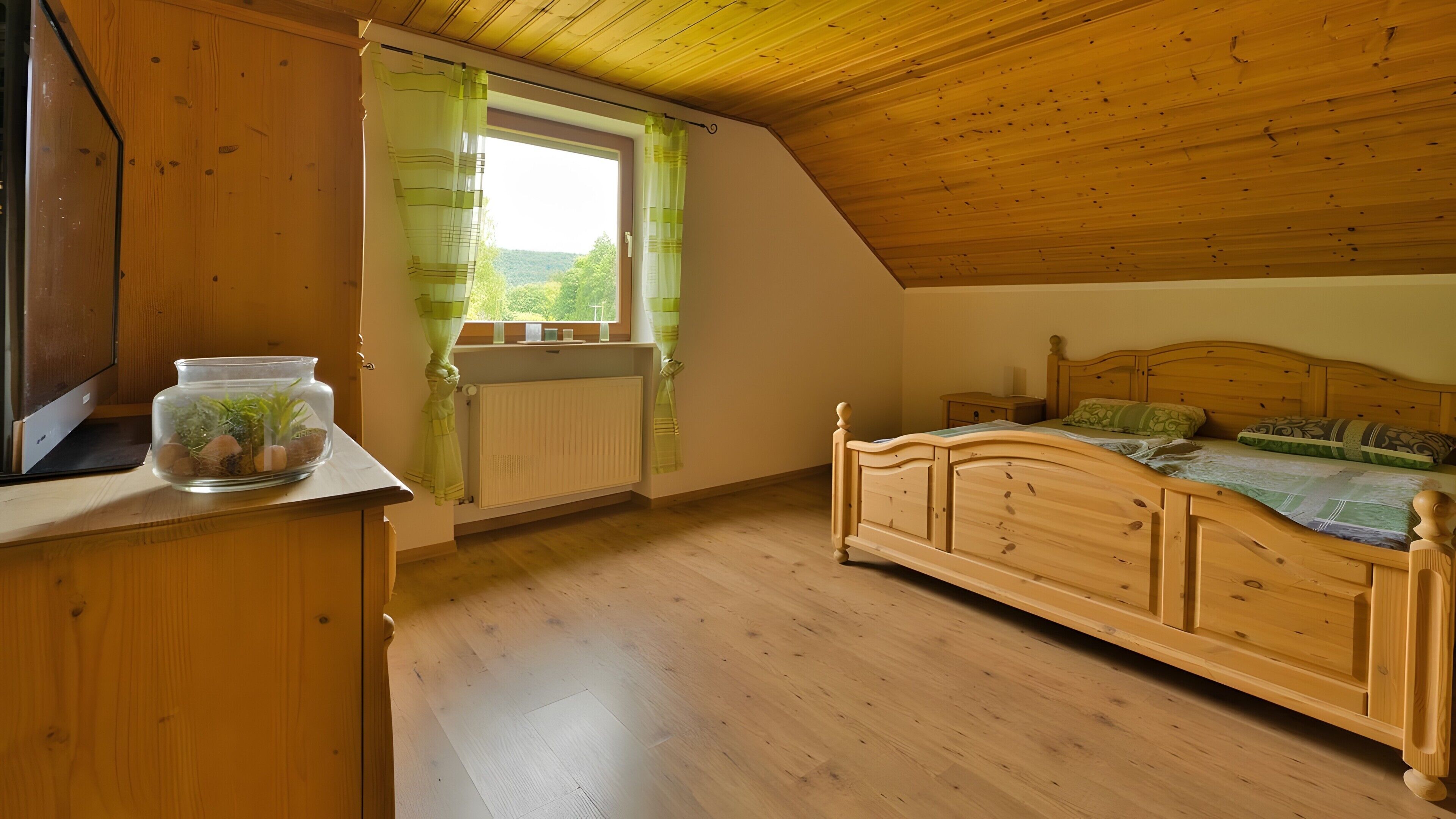 A bedroom with a wooden ceiling, a bed with a headboard, a window with curtains, a radiator, and a desk with a plant.