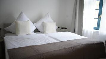 Ein Bett mit weißer Bettwäsche und zwei Kissen, ein Nachttisch, eine Steckdose und ein Fenster mit Vorhängen.