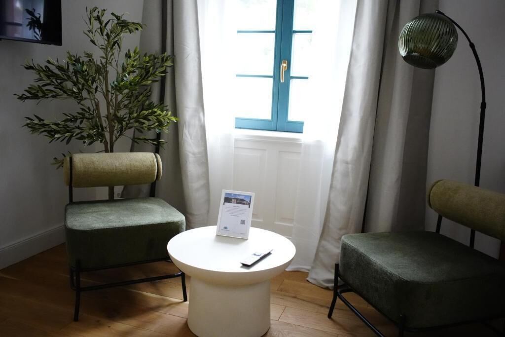 A room with two green chairs, a white table with a sign, a plant, a window with blue shutters, and a floor lamp.