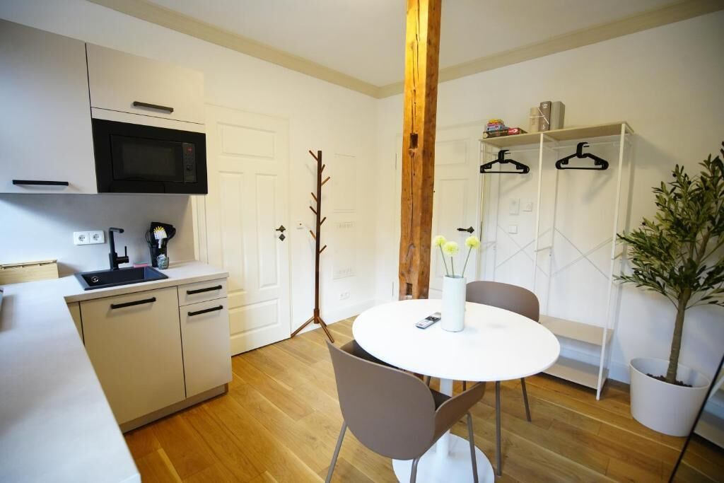 A compact kitchen with a microwave, sink, and cabinets, a round dining table with chairs, and a potted plant.