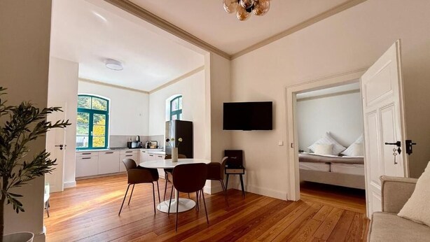 An open-plan living space with a dining area, a TV mounted on the wall, and a bed in the background. - Königsgleis-Apartments (Bad Wildbad)