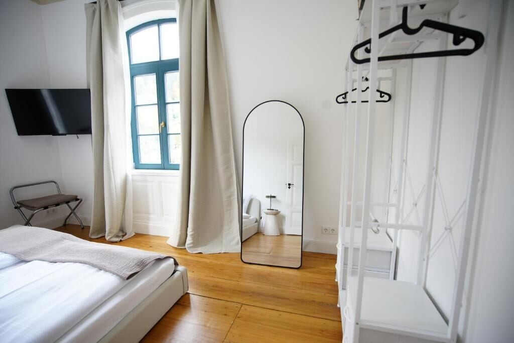 Un dormitorio con cama, televisor montado en la pared, ventana con cortinas, espejo y armario con perchero.
